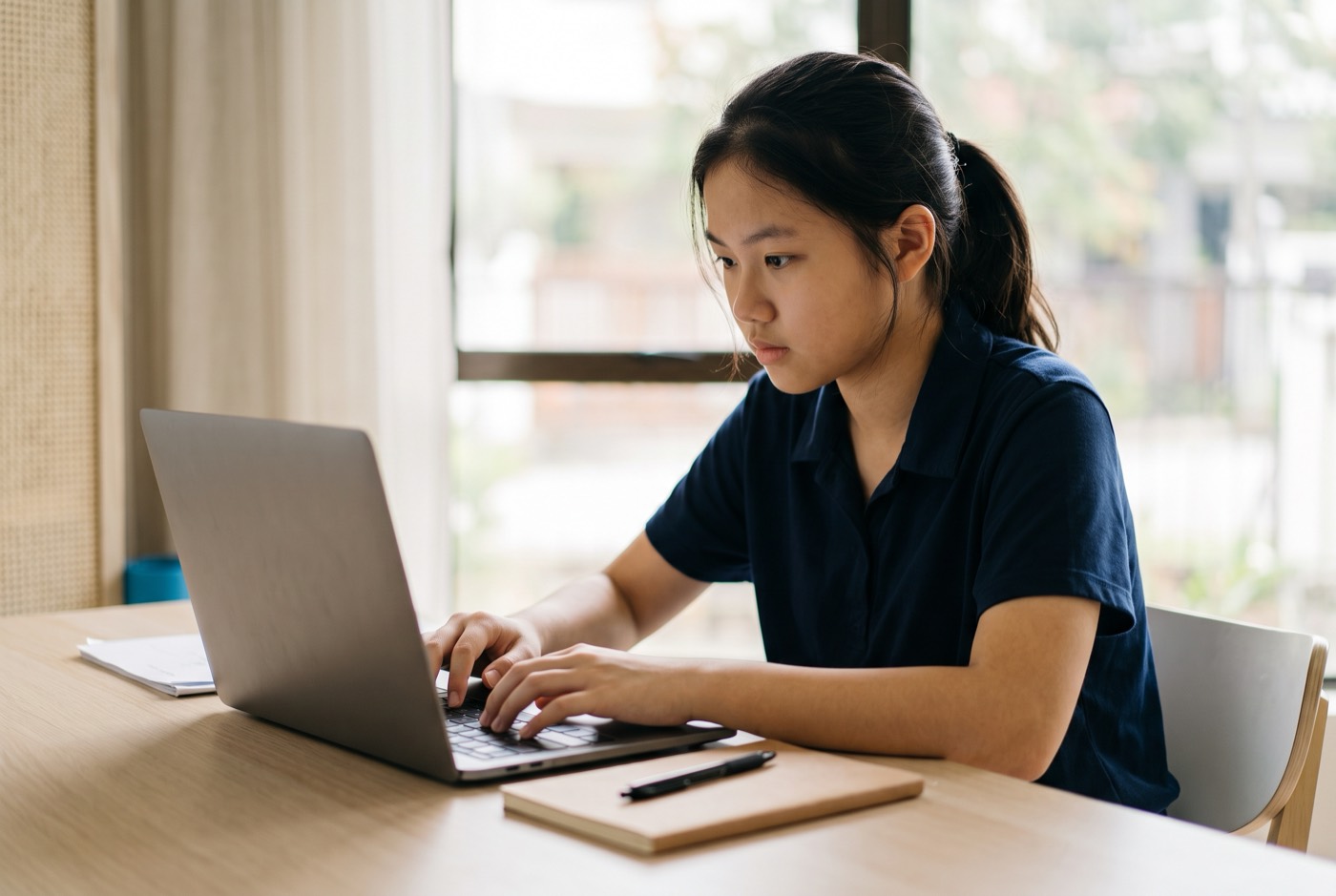 A high school student sitting at a clean desk with a laptop, displaying a web application build log alongside a unive...