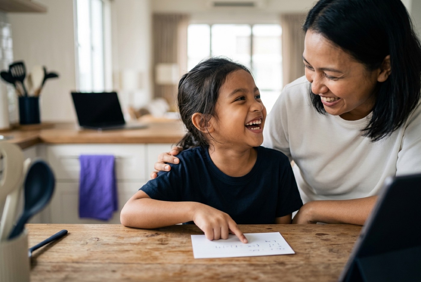 A young girl aged eight pointing at an index card on a wooden kitchen counter while laughing with her mother, indoor ...