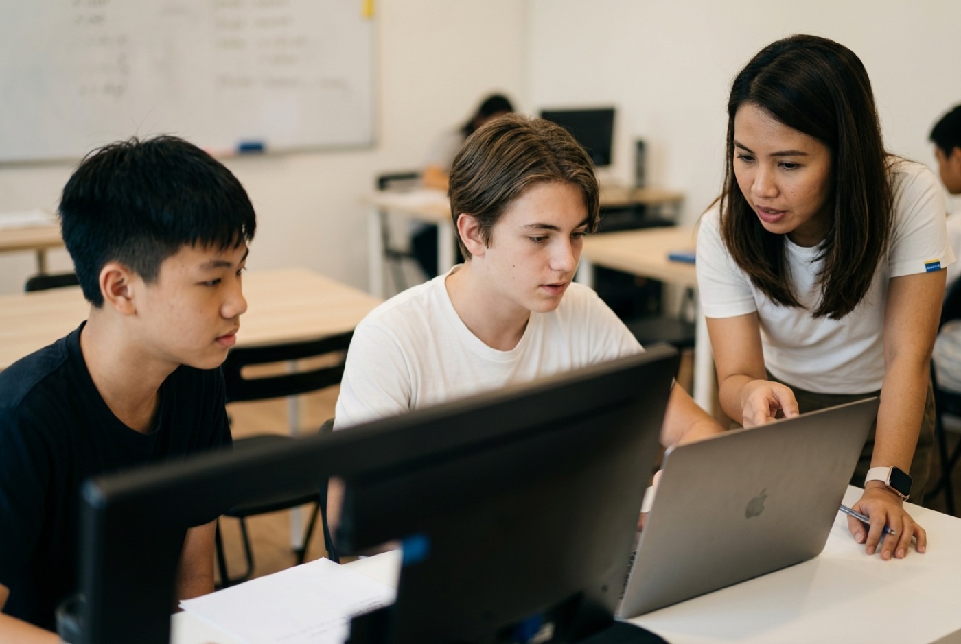 A teacher guiding two teenage students at a computer workstation as they review code and analytics graphs together in...